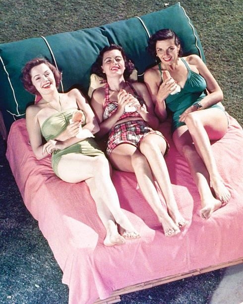 Jane Russell , American actress, with two other women, each wearing a swimsuit, smiling as they hold hotdogs on a sun lounger, circa 1950.