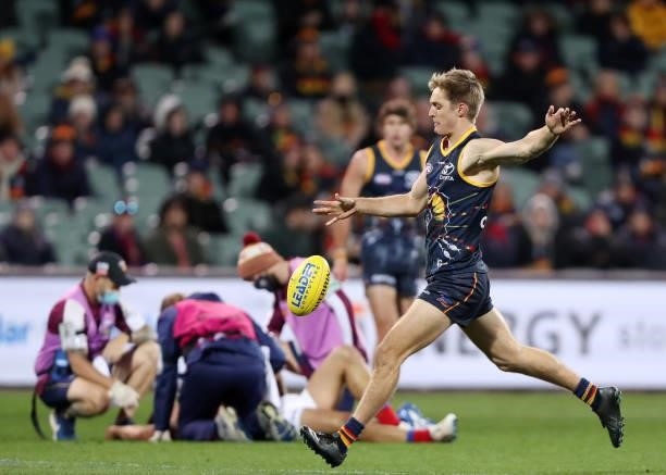 Jackson Hately of the Crows kicks the ball while Brandon Starcevich of the Lions his in the hands of trainers during the 2021 AFL Round 16 match...
