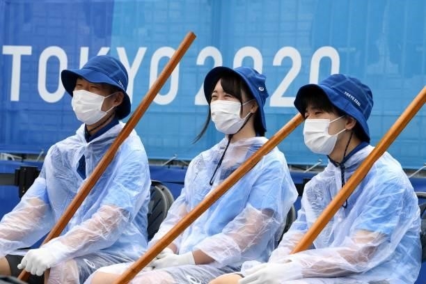 Attendants wearing face masks and ponchos sit with their rakes in the women's preliminary beach volleyball pool B match between the USA and Spain...