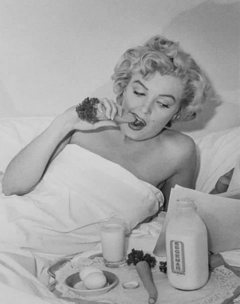 American actress and model Marilyn Monroe , partly covered by a sheet, holds a carrot between her teeth as she reads a script in bed at the Hotel...