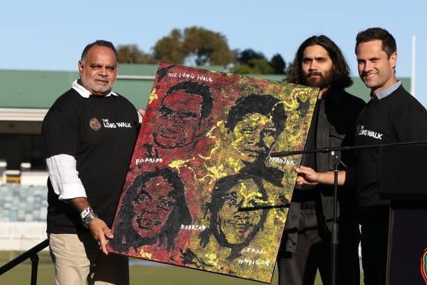 Michael Long and Gavin Wanganeen pose with artist Kambarni and his painting prior to the Long Walk from the WACA to Optus Stadium during the round 12...