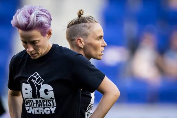 Megan Rapinoe of OL Reign with her head down wears a tee shirt that says Big Anti-Racist Energy with a Black Power Fist next to Jessica Fishlock of...
