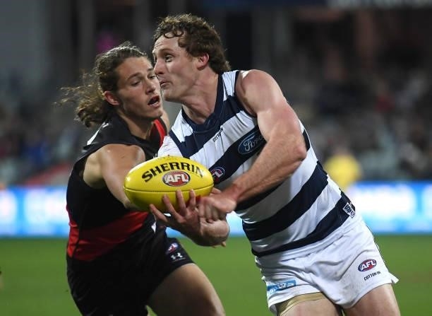Jed Bews of the Cats handballs whilst being tackled by Archie Perkins of the Bombers during the round 16 AFL match between Geelong Cats and Essendon...