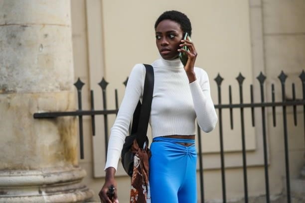 Model wears a white ribbed turtleneck crop-top, blue belted flared pants, a black shiny leather shoulder bag, a brown / black / white / orange print...