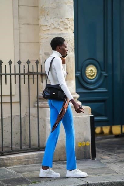 Model wears a white ribbed turtleneck crop-top, blue belted flared pants, a black shiny leather shoulder bag, a brown / black / white / orange print...