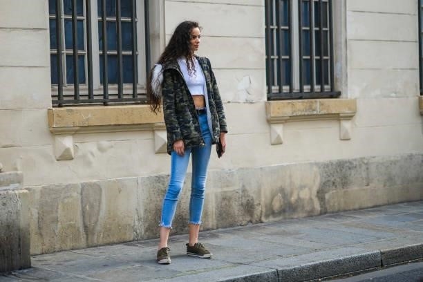 Model wears a white crop-top, a khaki brown and green military print pattern hoodie with white fur long puffer jacket, blue faded denim jeans ripped...