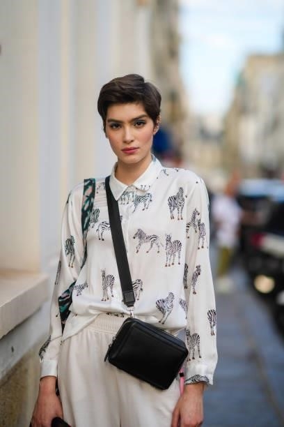 Model wears a white with black and white zebra animals patterns shirt, a black shiny leather crossbody bag, white flowing pants, a black with green...