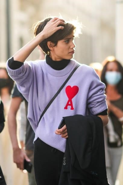 Model wears a black turtleneck pullover, a pale purple ribbed short sleeves pullover with a red A with heart embroidered, black pants, black shiny...