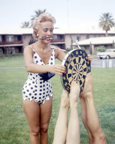 American singer, dancer and actress Jane Powell playing darts in a black and white, polka-dot, one-piece swimsuit, circa 1955. Her dartboard is being...